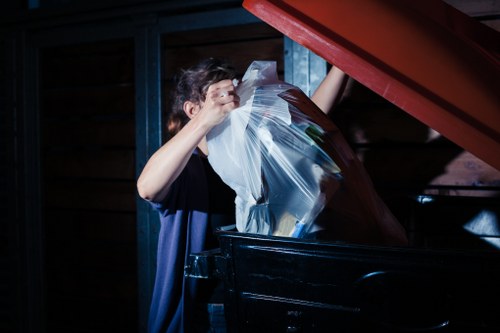 A person placing clear plastic bags filled with mixed domestic waste into a large black wheelie bin, situated outdoors on a paved area next to a dark wooden fence and blue metal support beams, with the lid of the bin open and the person's hand lifting the bag. The scene is illuminated by natural daylight, and the setting appears to be part of a residential or commercial premises in Dulwich, relevant to rubbish removal services provided by Commercial Waste Removal Dulwich. The person’s face is partially obscured, and they are wearing a dark shirt, with the focus on the detailed textures of the plastic bags and the metallic surface of the bin, emphasizing the collection of household waste materials.
