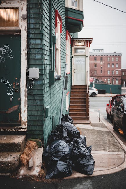 A residential sidewalk scene featuring a row of black wheelie bins with white painted numbers placed along the curbside in front of a brick terraced house. The bins are lined up closely, with some lid