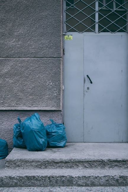 A large pile of black plastic rubbish bags and flattened cardboard boxes stacked against a beige, textured exterior wall in front of a red metal door. The door features graffiti tags in white and pink