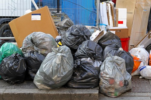 A collection of black, grey, and clear plastic rubbish bags filled with various waste materials, placed on a raised concrete surface outdoors, likely on a pavement or driveway. The bags exhibit different levels of fullness and are piled together, some tied at the top. Behind the bags, there are some cardboard boxes, a red container, and a few white and brown items, suggesting a mixture of household or commercial waste awaiting collection. Surrounding the scene, there are urban elements such as a metal fencing and building structures, indicating an area in Dulwich or nearby South London. The environment appears to be a typical street or alleyway setup where waste disposal is temporarily staged before pickup, aligning with rubbish removal services handled by companies like Commercial Waste Removal Dulwich. The lighting is natural, with a diffuse brightness hinting at an overcast day, ensuring all details such as material textures and colors are clearly visible for an accurate description geared towards accessibility and local SEO considerations.