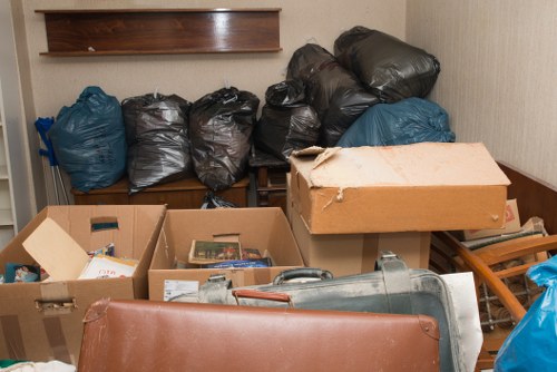 The image shows a cluttered indoor space, possibly a storage room or corner of a property, with multiple black and blue rubbish bags piled against a wooden shelf on the back wall. The bags are tightly packed and appear to contain mixed waste materials. In the foreground, there are open cardboard boxes filled with various items, including papers and small objects, positioned on a wooden surface or table. An orange-brown leather or faux leather chair is partially visible at the bottom left corner, with a metal object resembling a piece of equipment or debris lying nearby. The setting has neutral-colored walls and a wooden or laminate floor, with natural or indoor lighting illuminating the scene. The cluttered environment and the presence of sealed waste bags indicate a rubbish removal task, which commercial waste removal services in Dulwich, such as those at commercialwasteremovaldulwich.co.uk, could efficiently manage, especially in densely populated or residential areas in nearby postcode districts.
